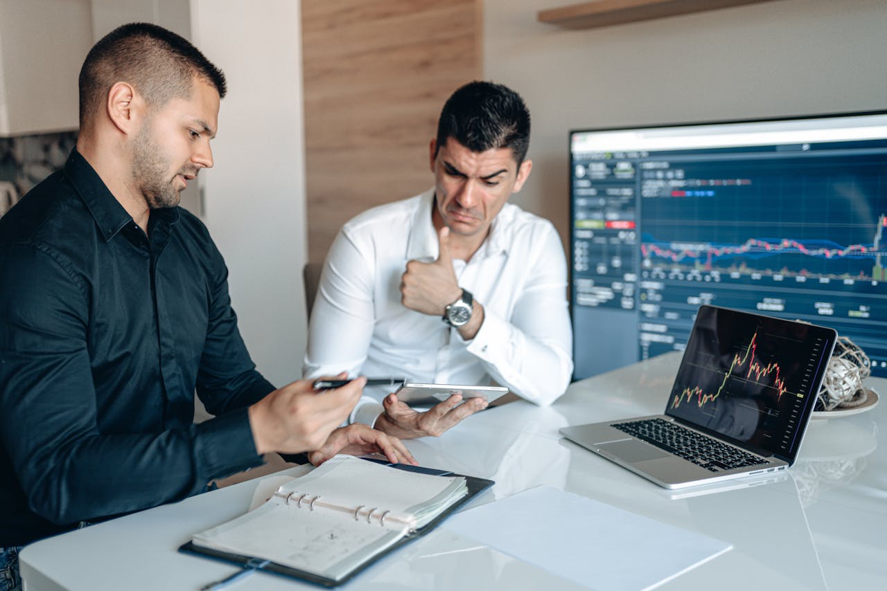 who-we-are Two professionals discuss data analysis on digital devices in an office setting, focusing on graphs and charts.